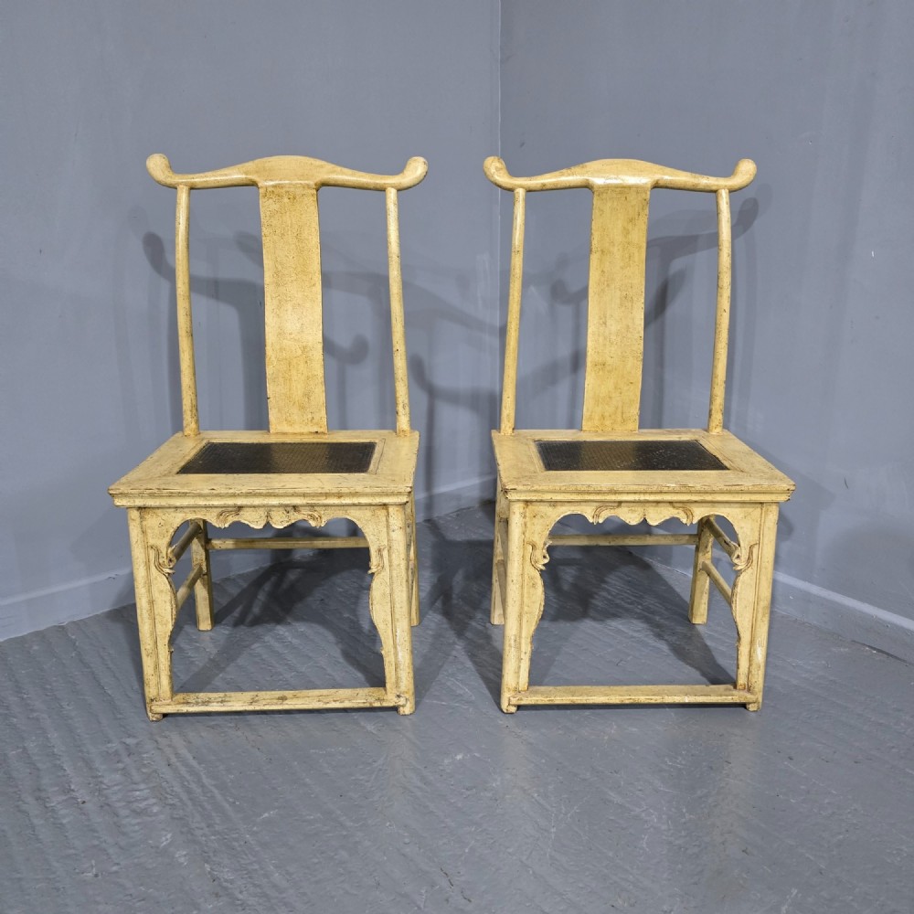 A pair of cream lacquered Chinese hall chairs featuring curved yoke crest rails, shaped splats, carved aprons, and dark inset seats.