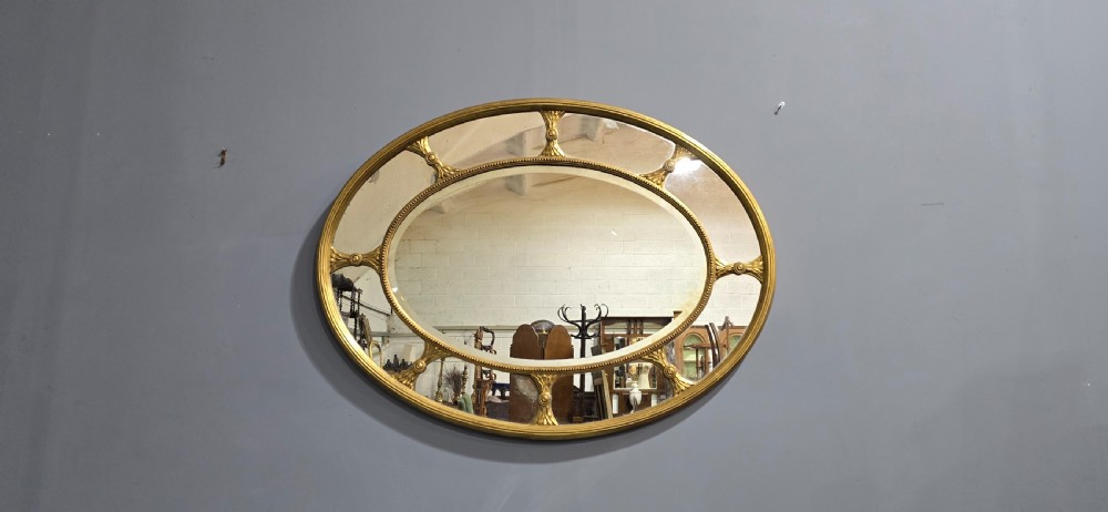 A large oval wall mirror with a beaded gold giltwood frame surrounding a central glass plate and outer sectional glass border. View II