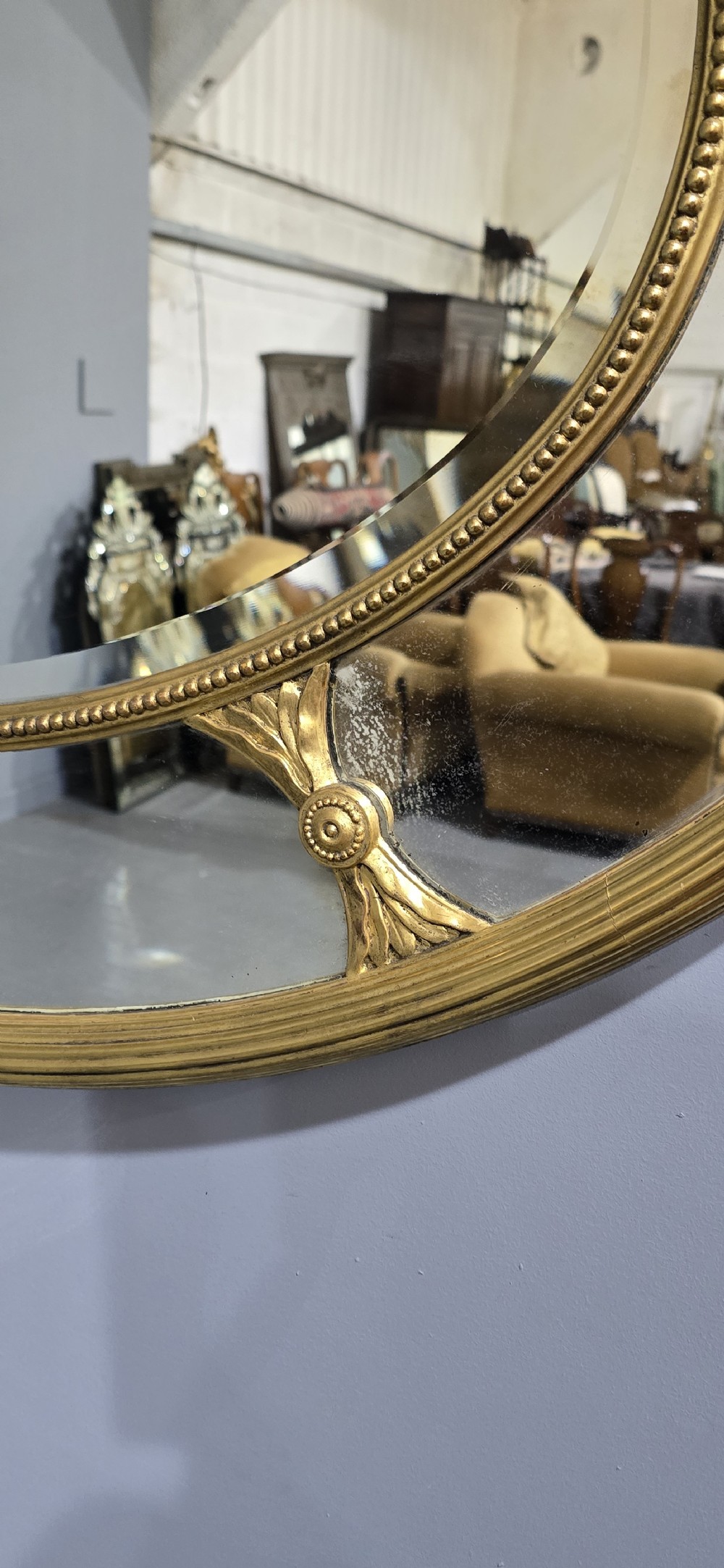 A large oval wall mirror with a beaded gold giltwood frame surrounding a central glass plate and outer sectional glass border. View IV