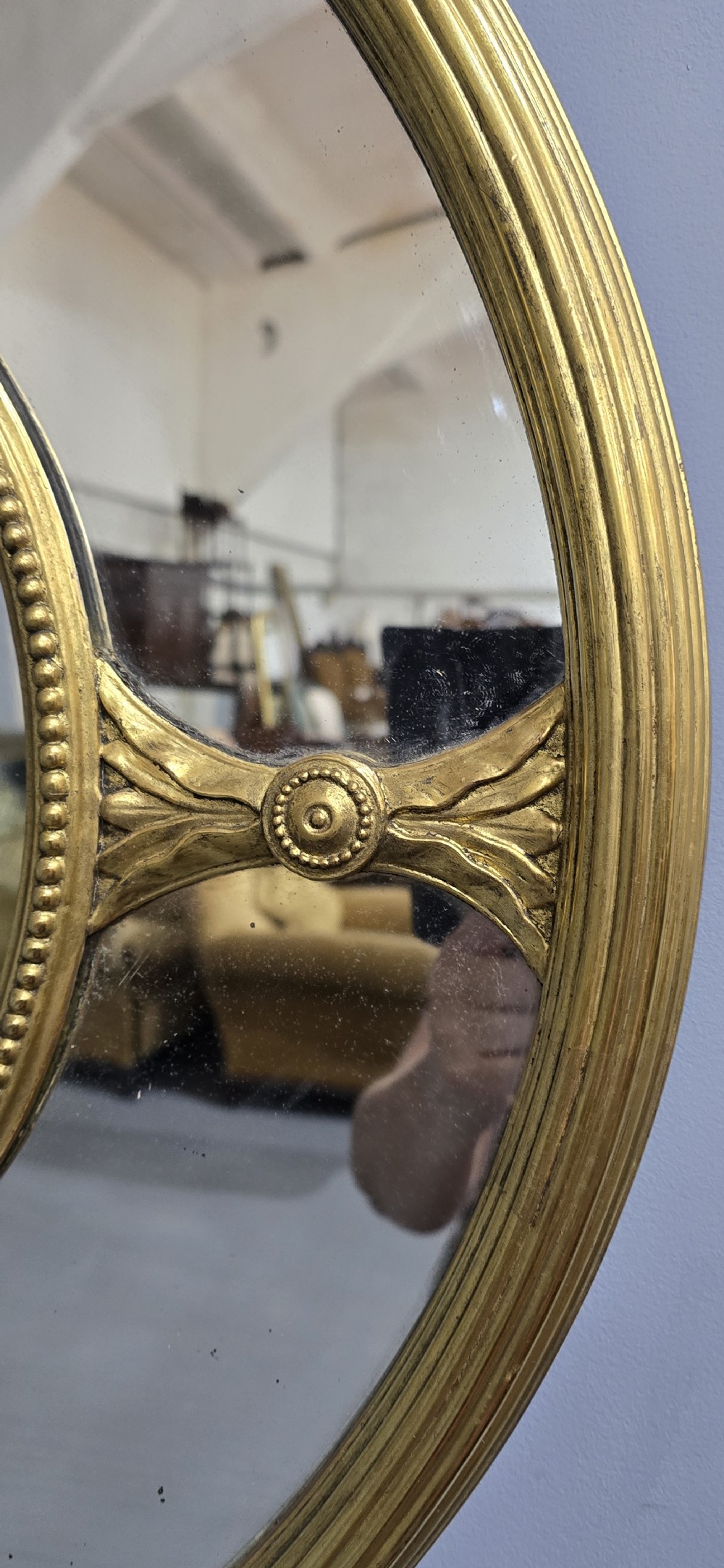 A large oval wall mirror with a beaded gold giltwood frame surrounding a central glass plate and outer sectional glass border. View V