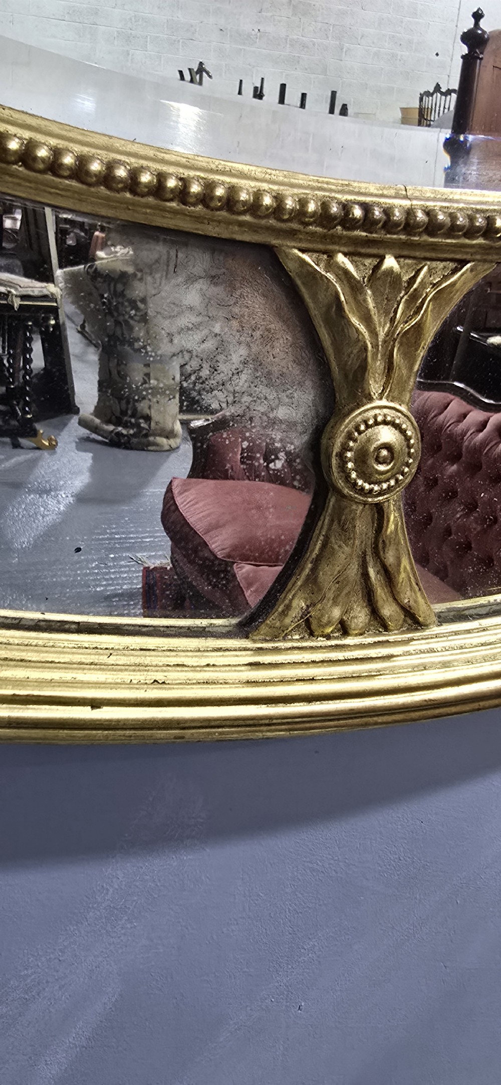 A large oval wall mirror with a beaded gold giltwood frame surrounding a central glass plate and outer sectional glass border. View VI