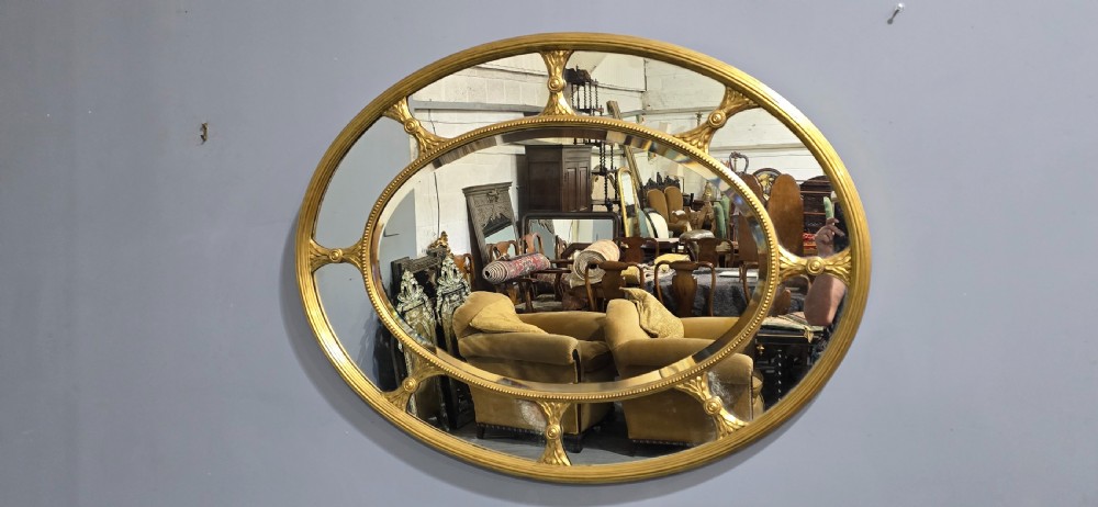 A large oval wall mirror with a beaded gold giltwood frame surrounding a central glass plate and outer sectional glass border. View VII