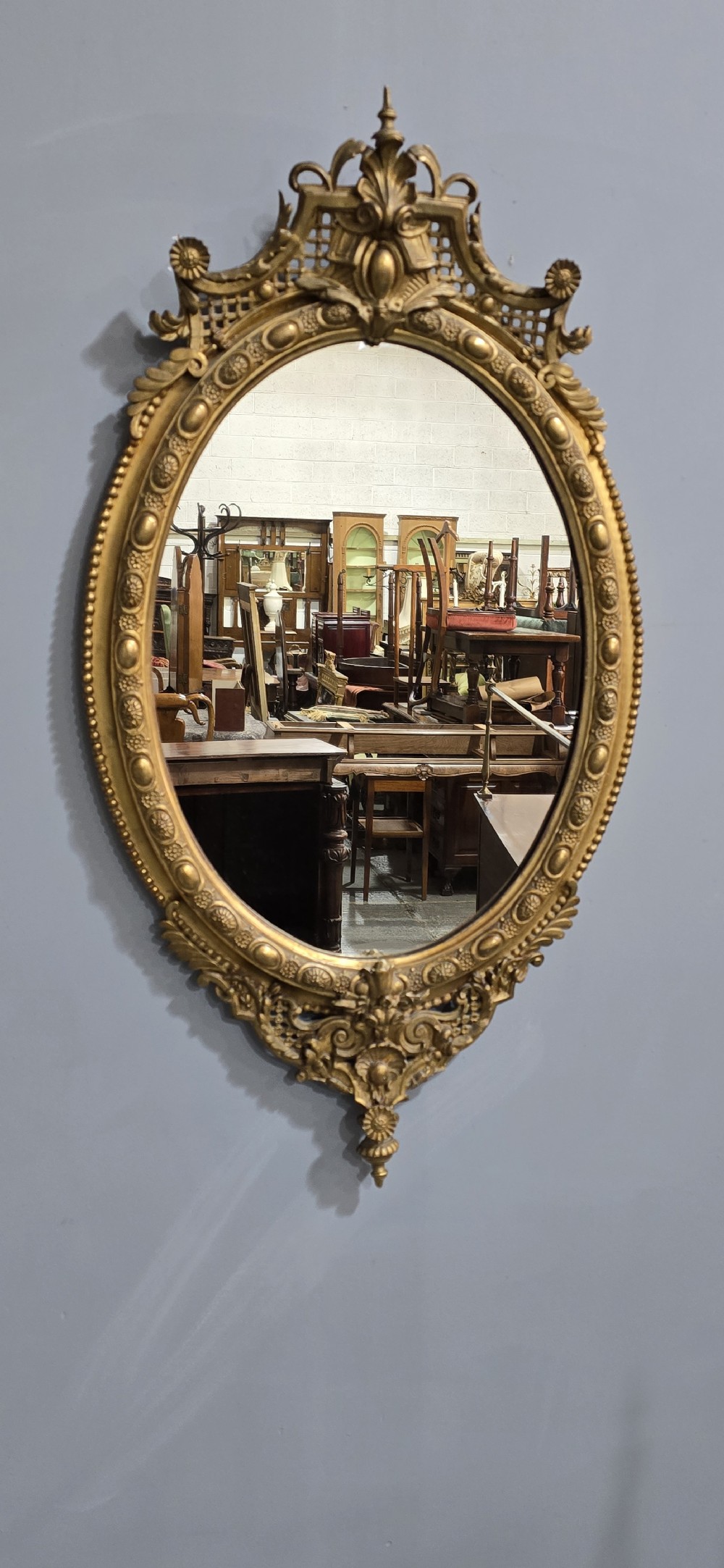 An oval giltwood wall mirror featuring an ornate carved crest with lattice detail, a beaded inner border, and a decorative bottom pendant. View IV