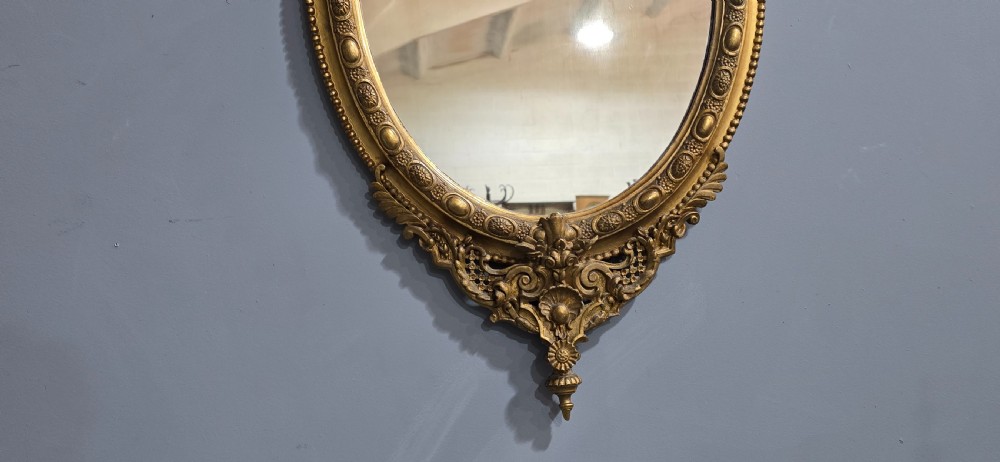 An oval giltwood wall mirror featuring an ornate carved crest with lattice detail, a beaded inner border, and a decorative bottom pendant. View V