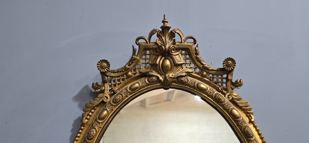 An oval giltwood wall mirror featuring an ornate carved crest with lattice detail, a beaded inner border, and a decorative bottom pendant. View VI