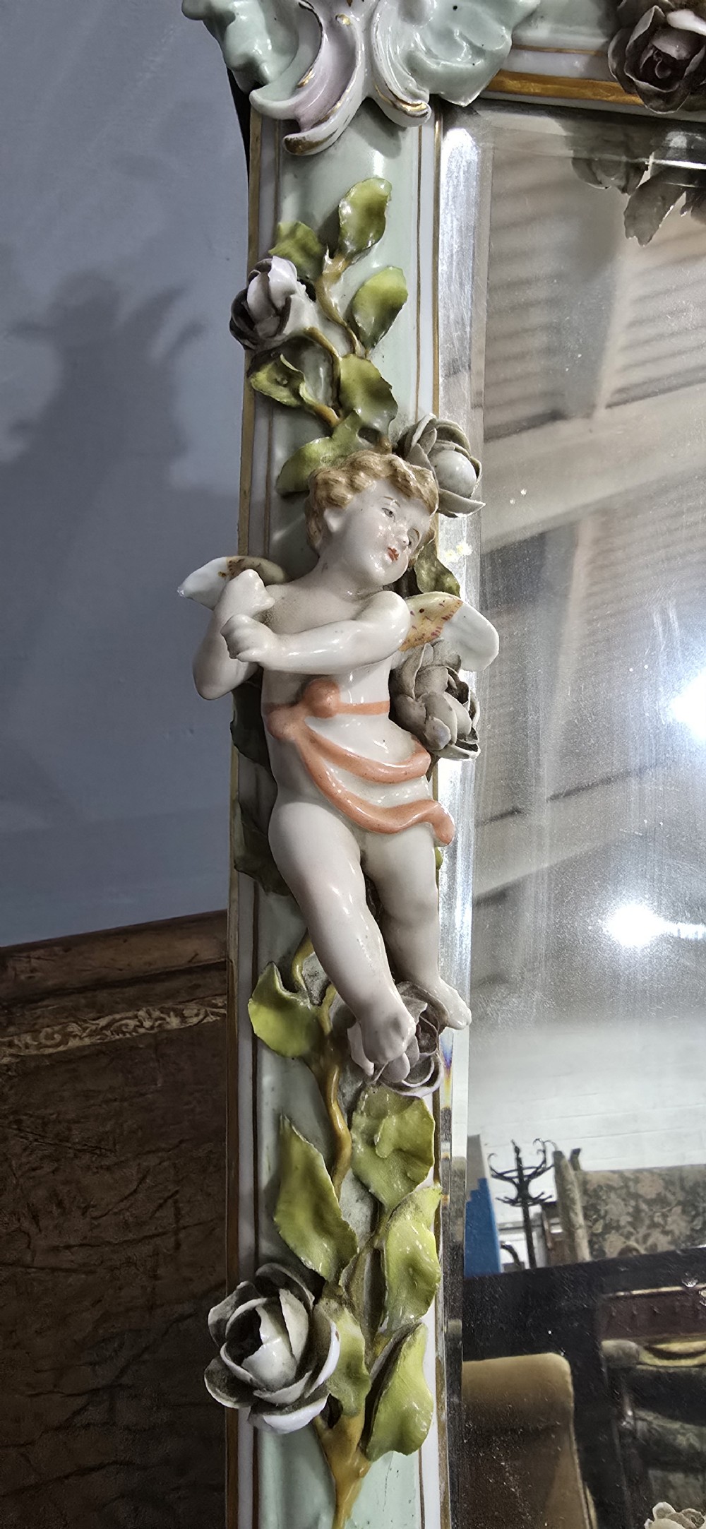 An antique Dresden porcelain easel mirror decorated with sculpted cherubs, encrusted pastel flowers, and Rococo scrollwork around a rectangular glass frame. View IV