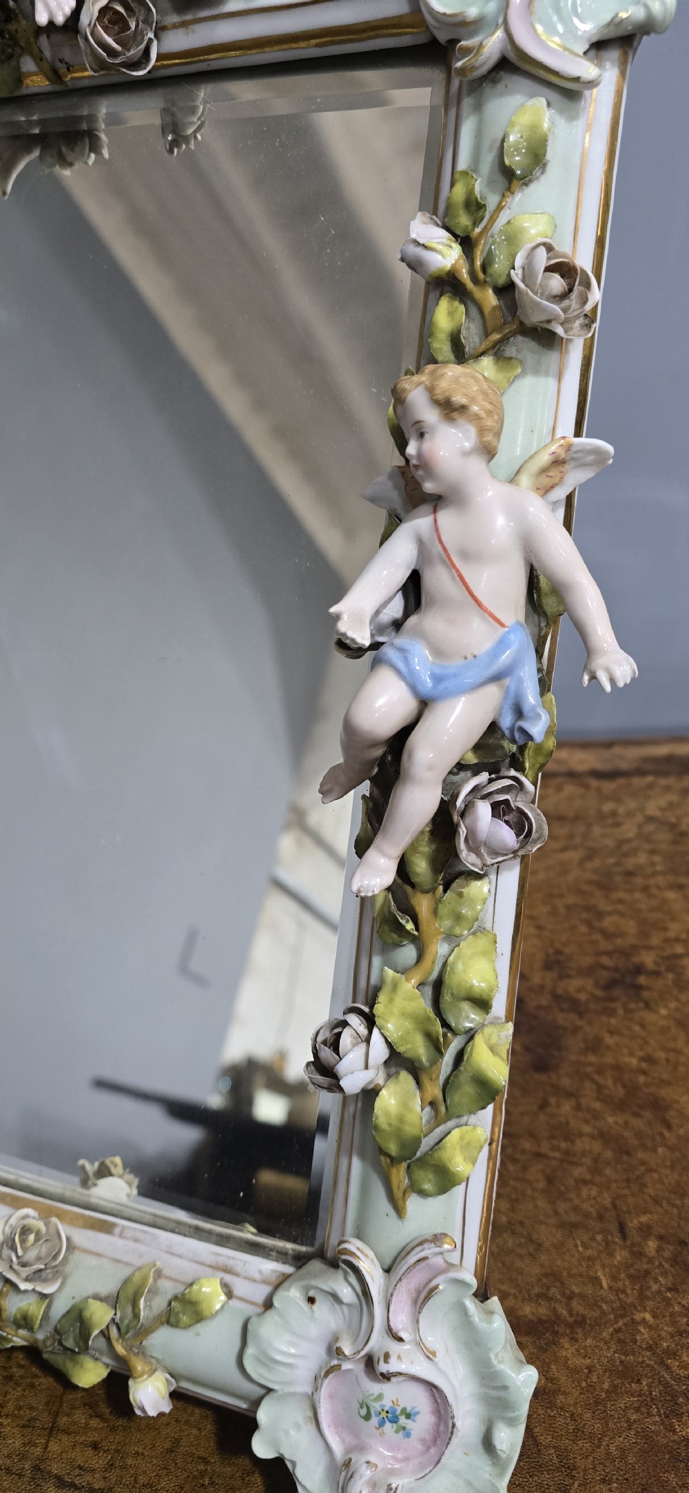 An antique Dresden porcelain easel mirror decorated with sculpted cherubs, encrusted pastel flowers, and Rococo scrollwork around a rectangular glass frame. View VI