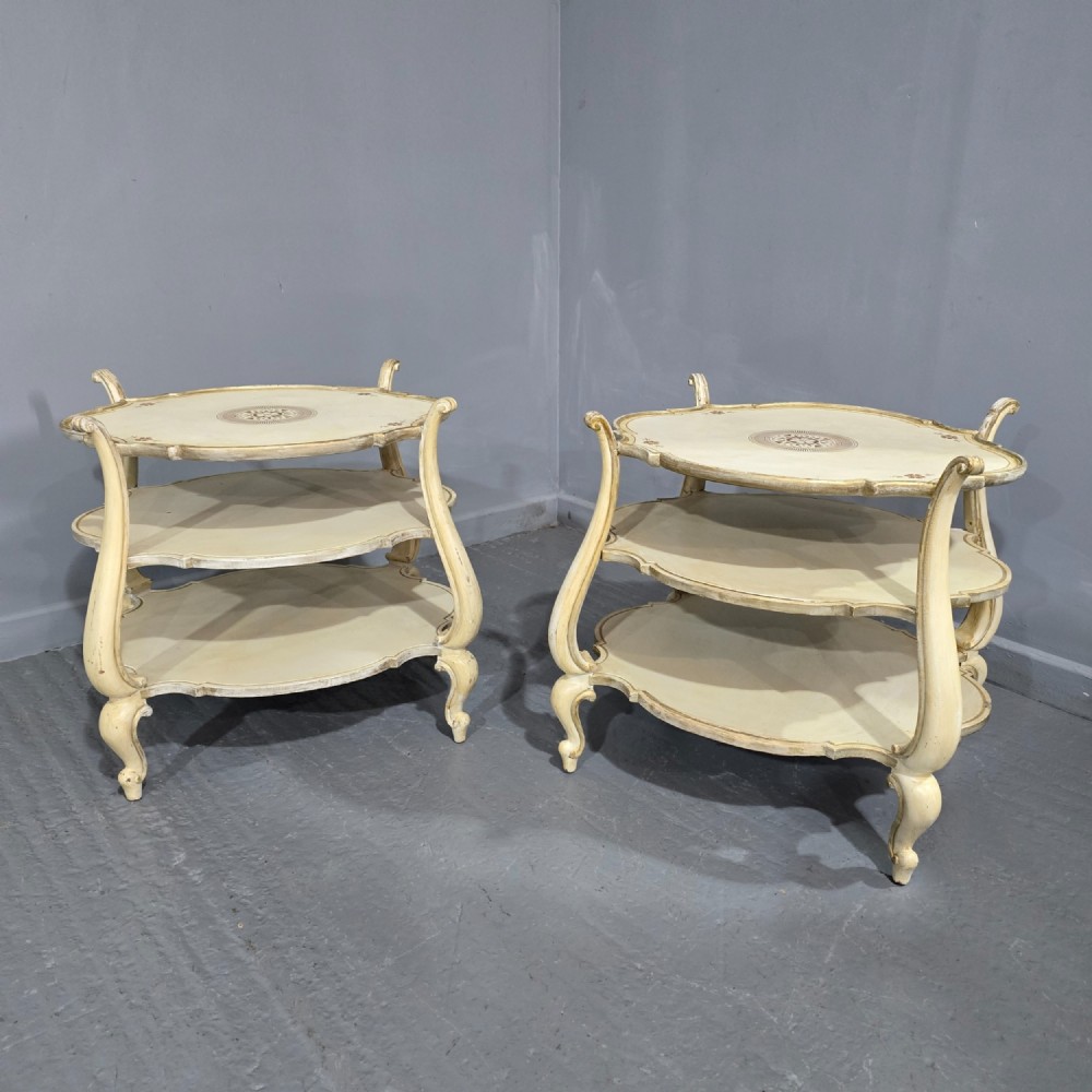 A pair of cream painted three-tier occasional lamp tables with cabriole legs and painted floral medallion tops.