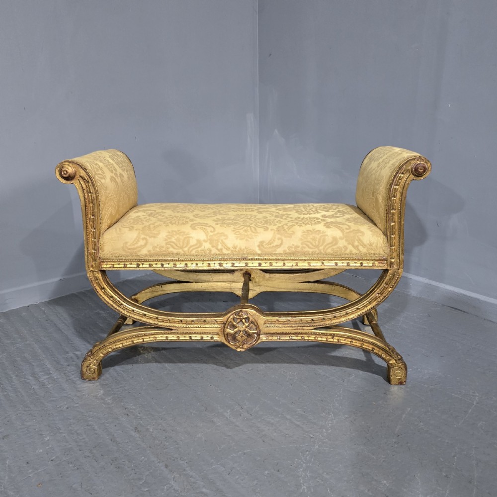 A French Neoclassical giltwood window seat featuring scrolled armrests, carved beaded frame details, and gold damask upholstery.