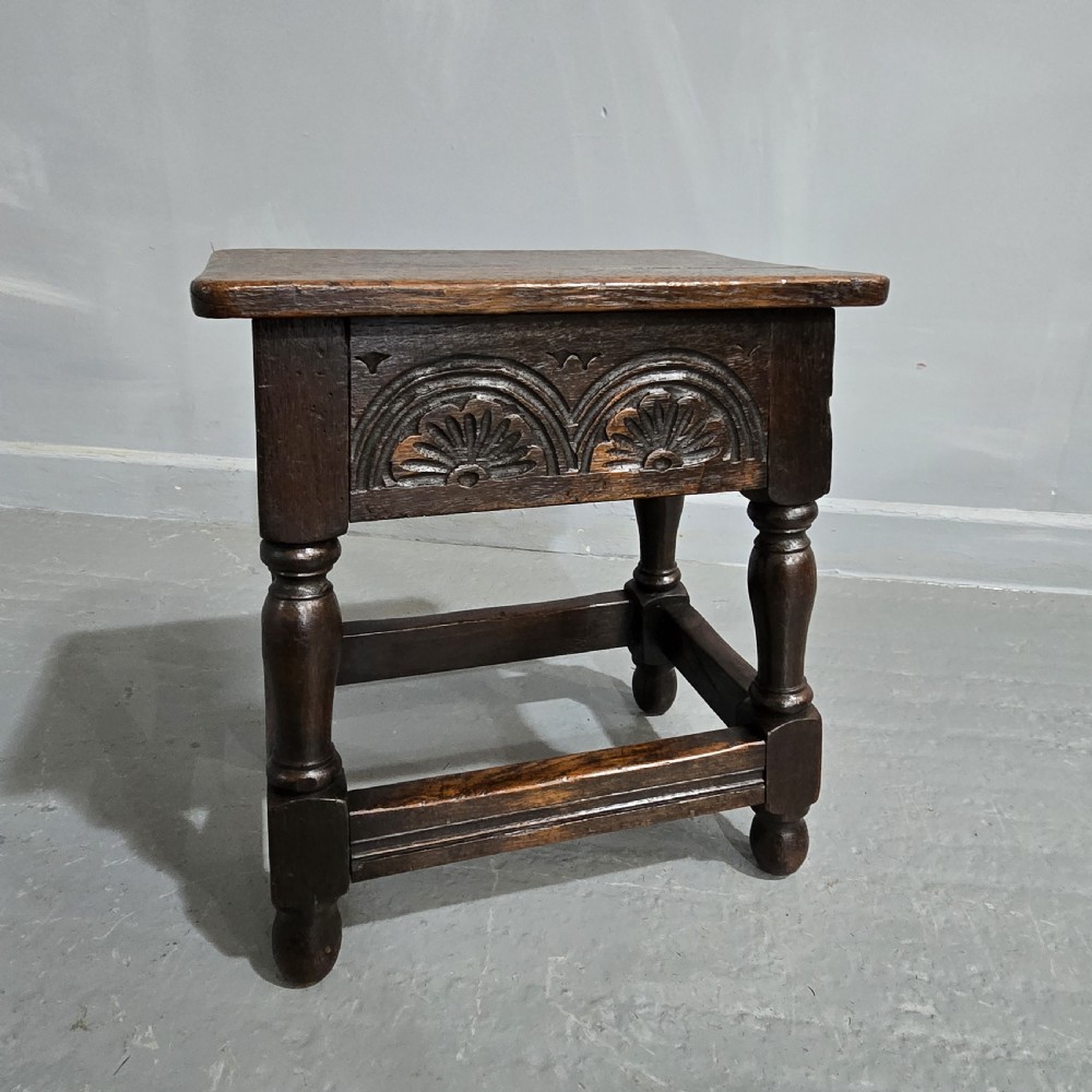 A dark oak joint stool featuring carved lunette detail along the frieze, turned legs, and lower wooden stretchers.