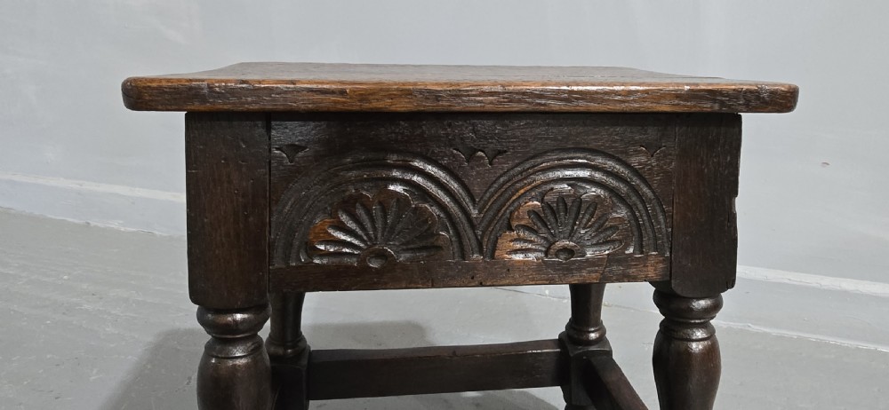 A dark oak joint stool featuring carved lunette detail along the frieze, turned legs, and lower wooden stretchers. View II