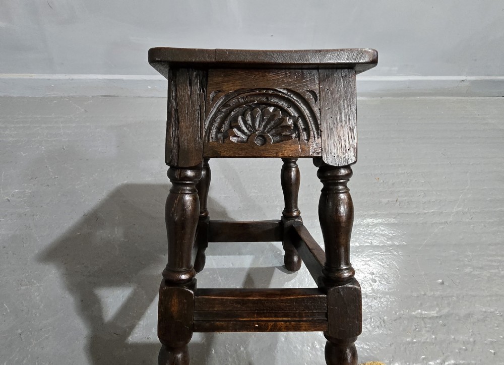 A dark oak joint stool featuring carved lunette detail along the frieze, turned legs, and lower wooden stretchers. View VI