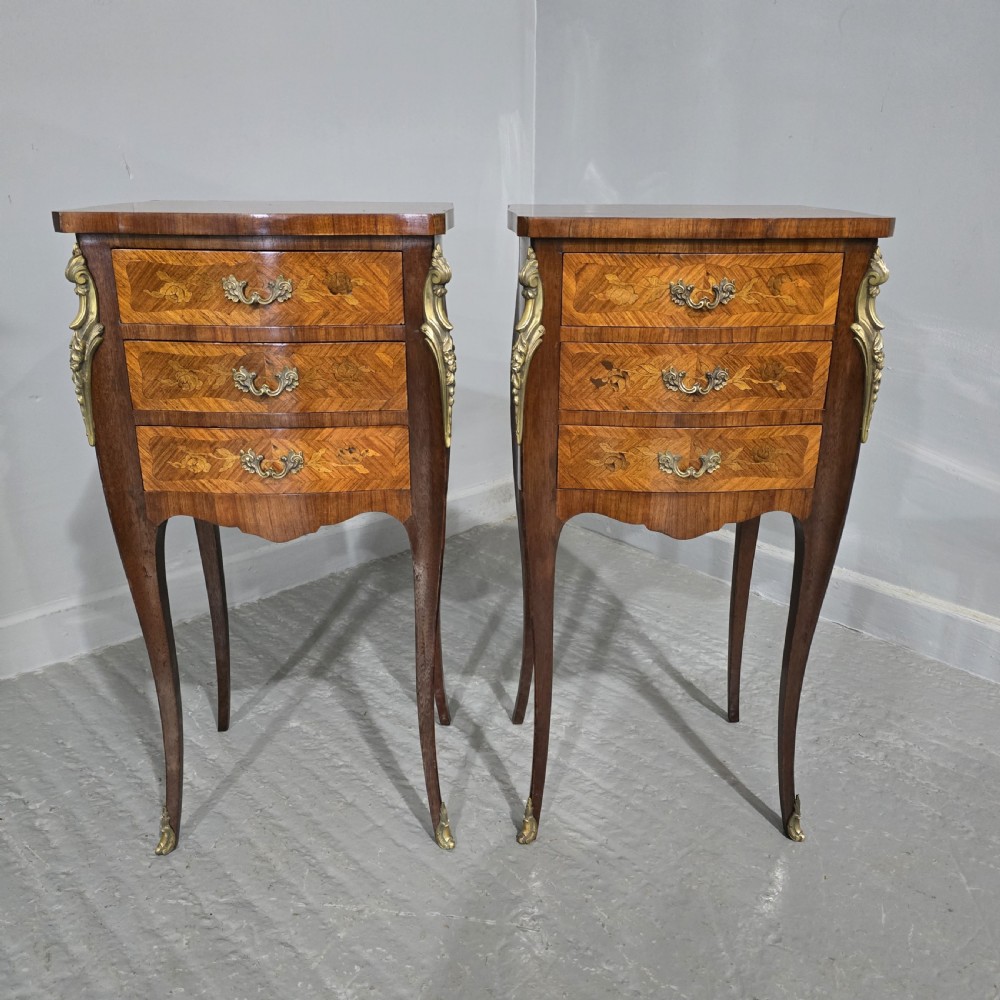 A pair of French Louis XV style bedside tables featuring floral marquetry wood veneers, three drawers, gilt brass mounts, and slender cabriole legs.