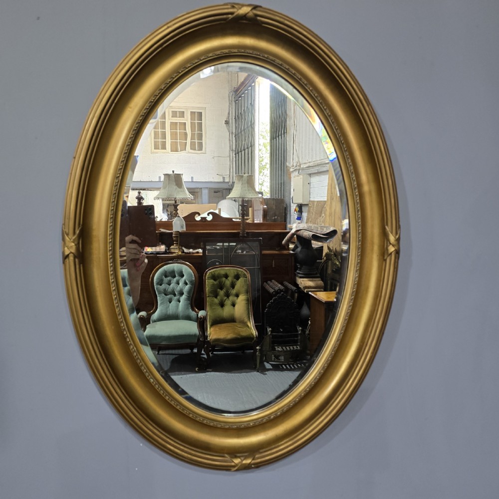 A large Victorian oval wall mirror featuring a ribbed gilt frame with crossed ribbon motifs surrounding a bevelled glass mirror plate.