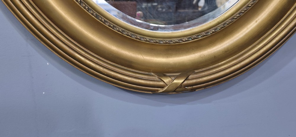 A large Victorian oval wall mirror featuring a ribbed gilt frame with crossed ribbon motifs surrounding a bevelled glass mirror plate. View V