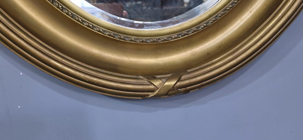 A large Victorian oval wall mirror featuring a ribbed gilt frame with crossed ribbon motifs surrounding a bevelled glass mirror plate. View VIII
