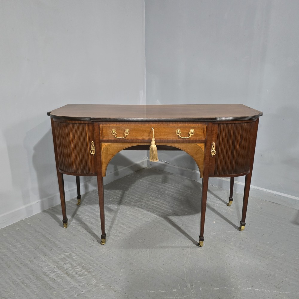A George III style inlaid mahogany sideboard featuring a central drawer above an arched aperture, flanked by vertical fluted cupboards on tapered legs with brass castors.