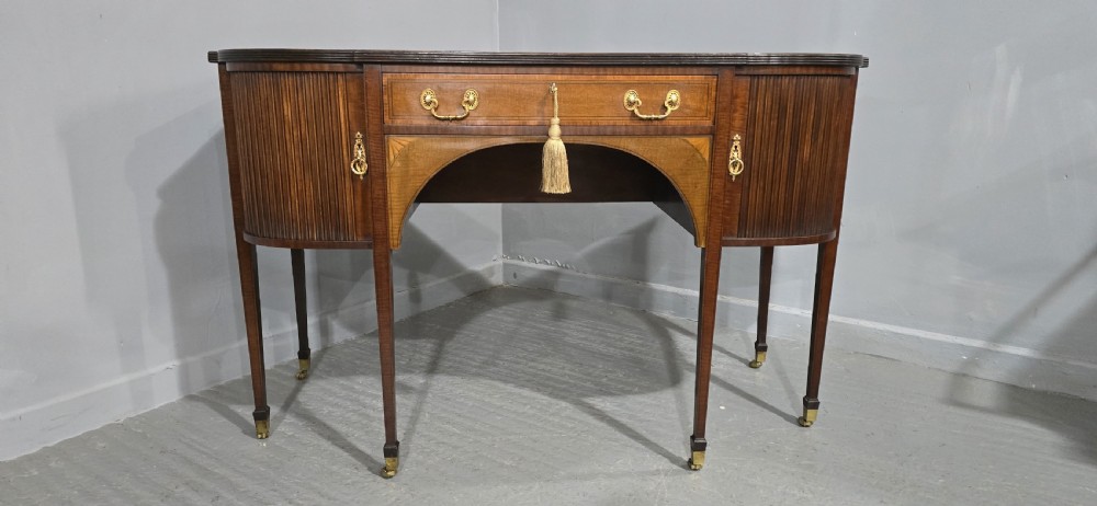 A George III style inlaid mahogany sideboard featuring a central drawer above an arched aperture, flanked by vertical fluted cupboards on tapered legs with brass castors. View II