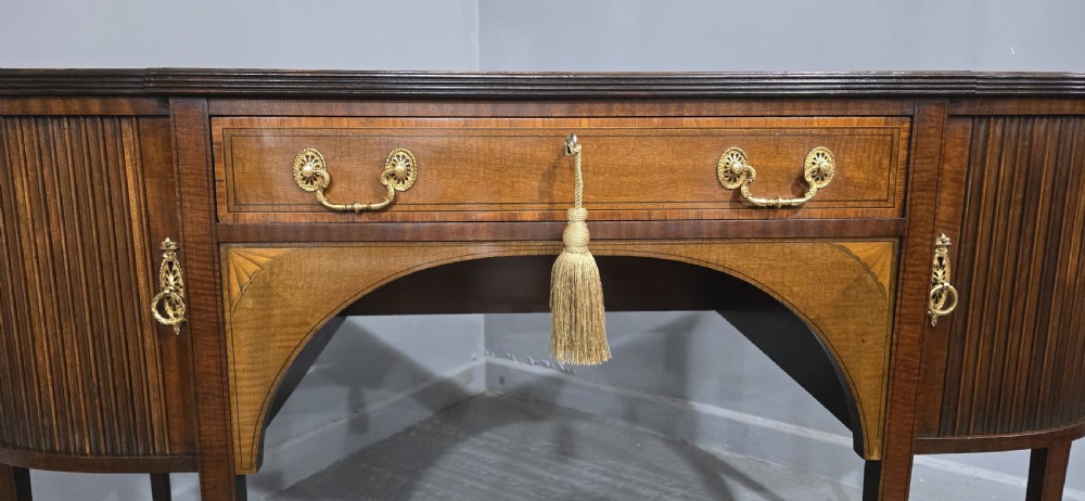 A George III style inlaid mahogany sideboard featuring a central drawer above an arched aperture, flanked by vertical fluted cupboards on tapered legs with brass castors. View V