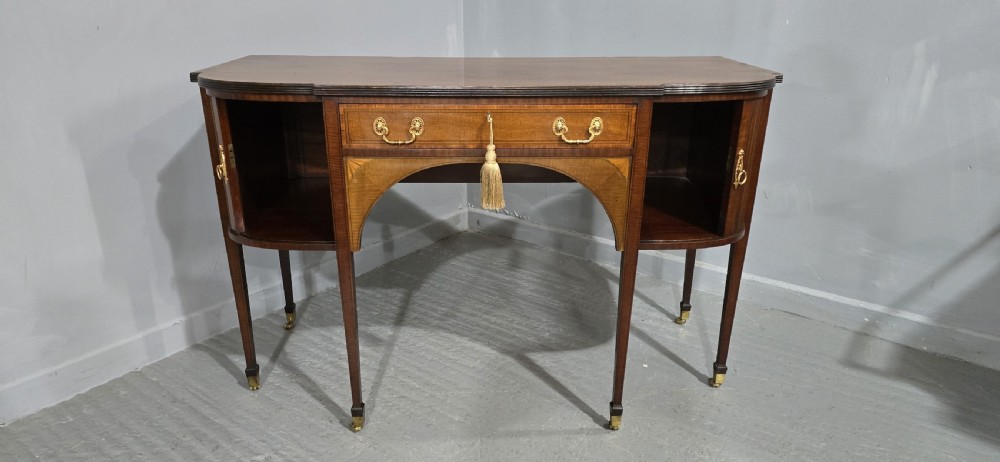 A George III style inlaid mahogany sideboard featuring a central drawer above an arched aperture, flanked by vertical fluted cupboards on tapered legs with brass castors. View VII
