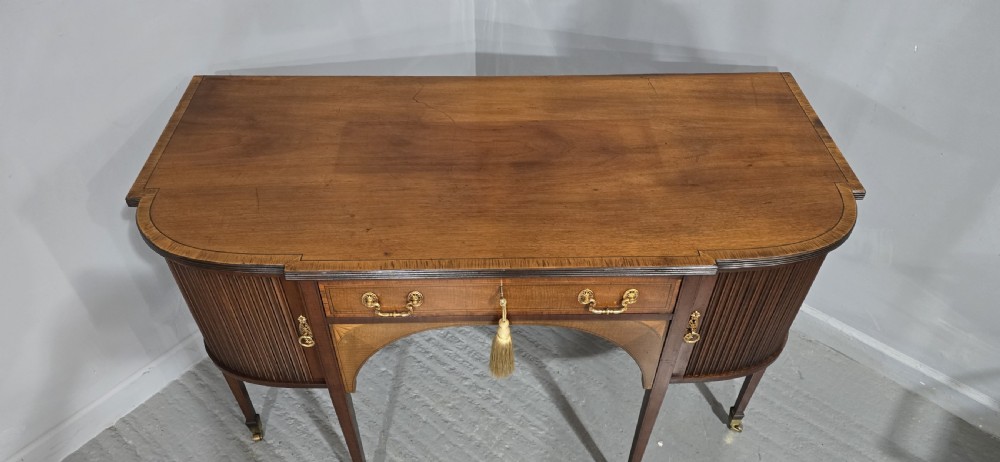 A George III style inlaid mahogany sideboard featuring a central drawer above an arched aperture, flanked by vertical fluted cupboards on tapered legs with brass castors. View X