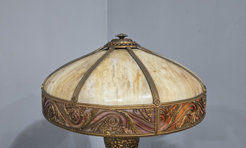 An Art Nouveau table lamp featuring a domed amber and red slag glass shade with decorative metal filigree set on a cast bronze floral base. View IV