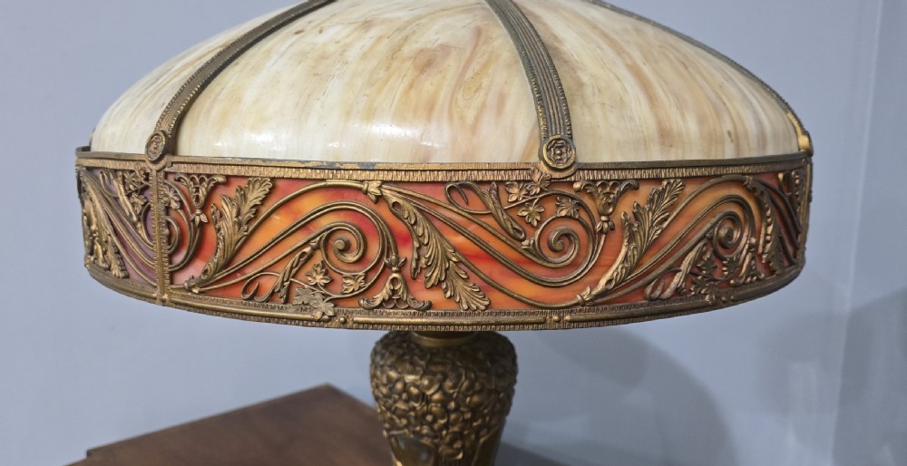 An Art Nouveau table lamp featuring a domed amber and red slag glass shade with decorative metal filigree set on a cast bronze floral base. View VI