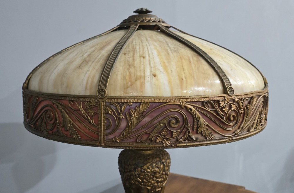 An Art Nouveau table lamp featuring a domed amber and red slag glass shade with decorative metal filigree set on a cast bronze floral base. View VII
