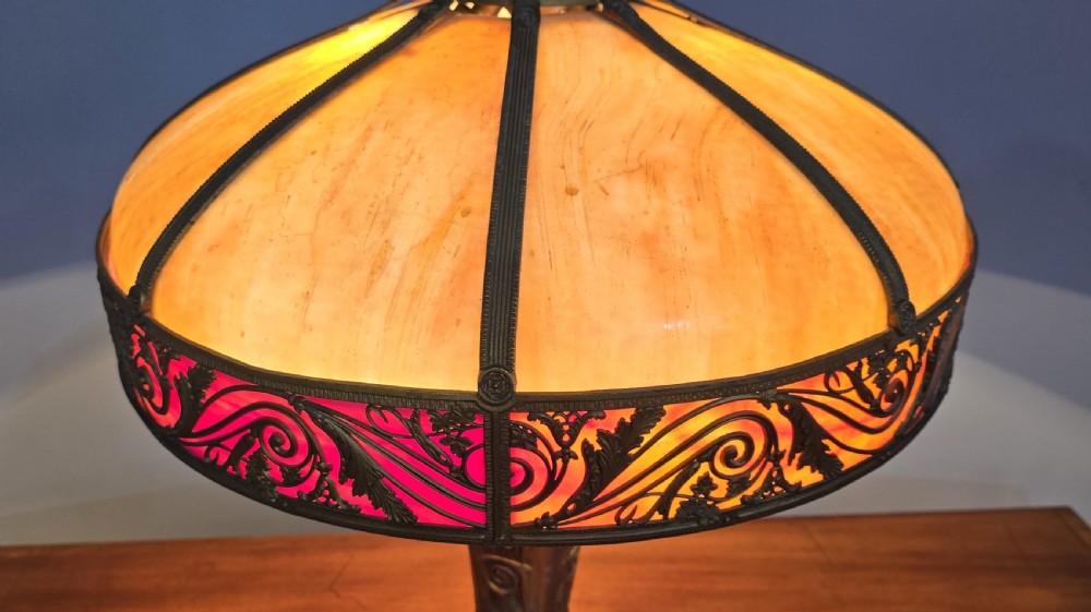 An Art Nouveau table lamp featuring a domed amber and red slag glass shade with decorative metal filigree set on a cast bronze floral base. View IX