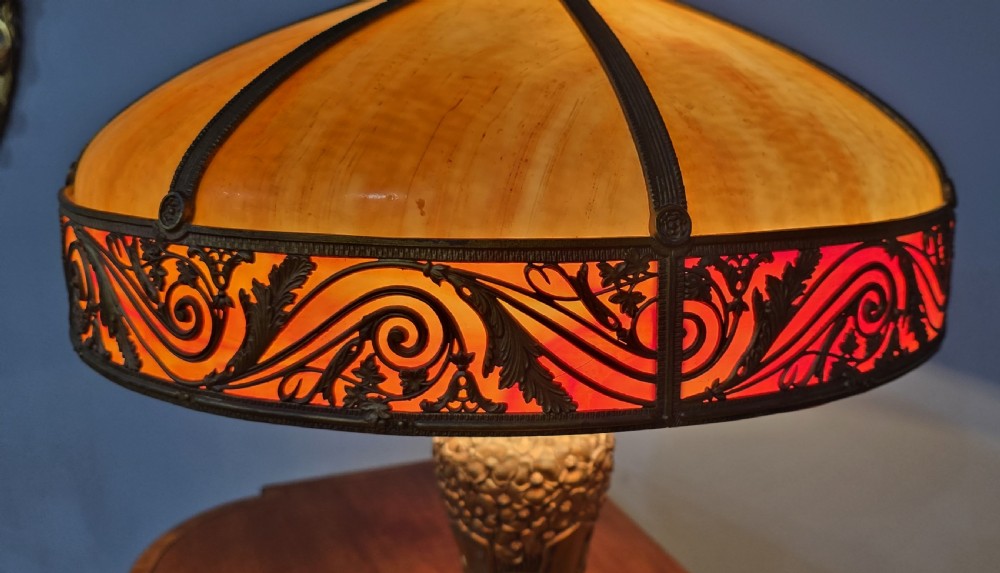 An Art Nouveau table lamp featuring a domed amber and red slag glass shade with decorative metal filigree set on a cast bronze floral base. View XI