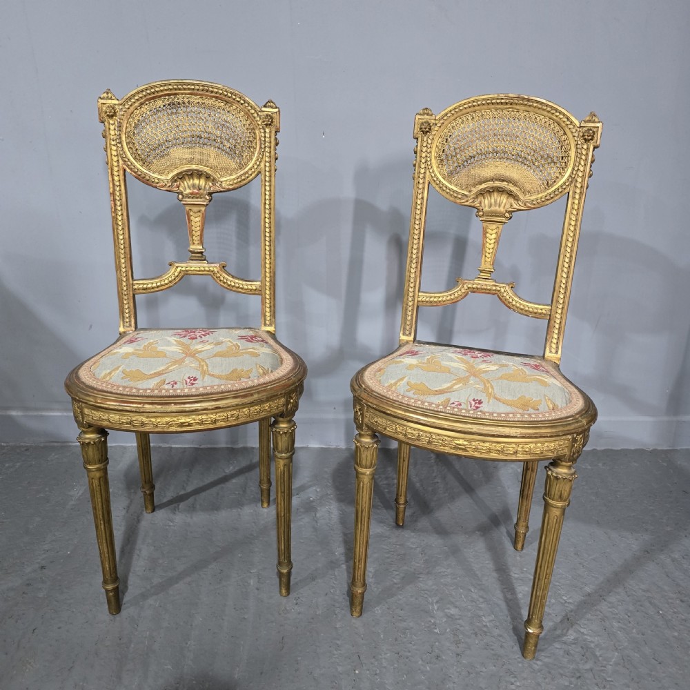 A pair of gold giltwood Louis XVI style side chairs featuring oval cane backs, carved splats, fluted legs, and patterned seat upholstery.