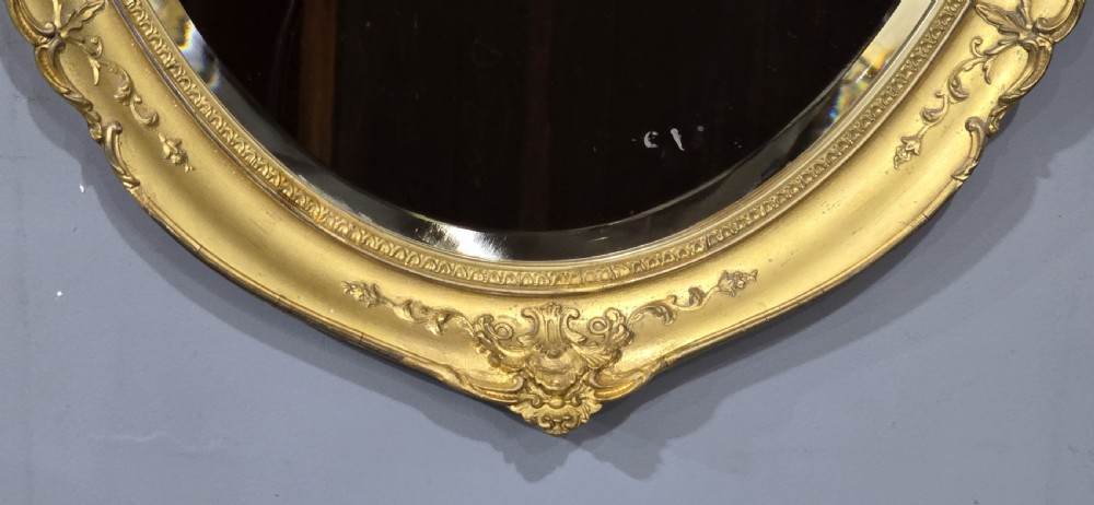 A vertical oval wall mirror featuring an ornate gold gilded frame decorated with scrolling floral motifs and bevelled glass. View IV