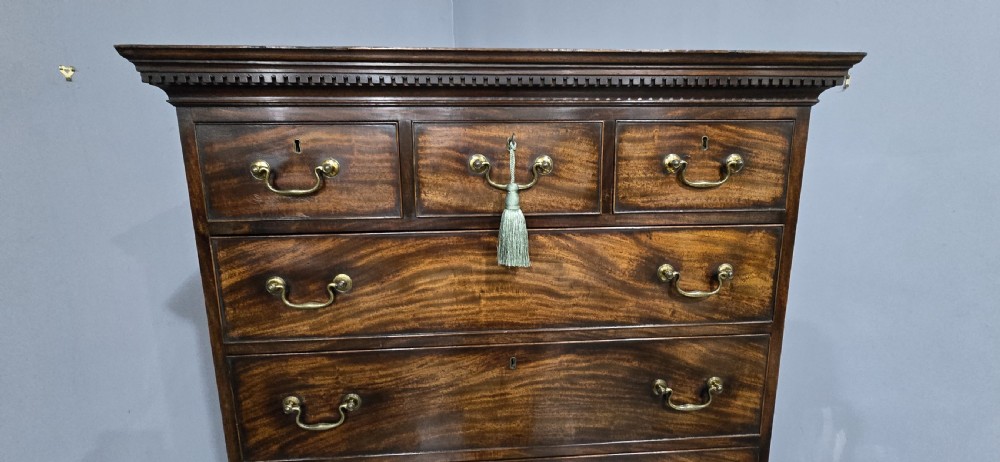 A tall Georgian mahogany chest on chest featuring two stacked drawer sections, flame mahogany grain, dentil cornice moulding, brass swan-neck handles, and bracket feet. View XI