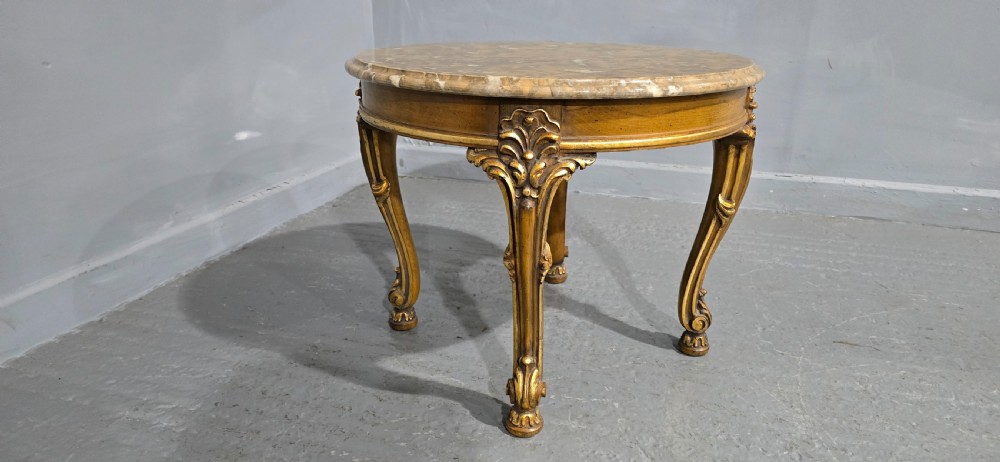 A circular French style low coffee table with a polished beige marble top and carved giltwood cabriole legs. View II
