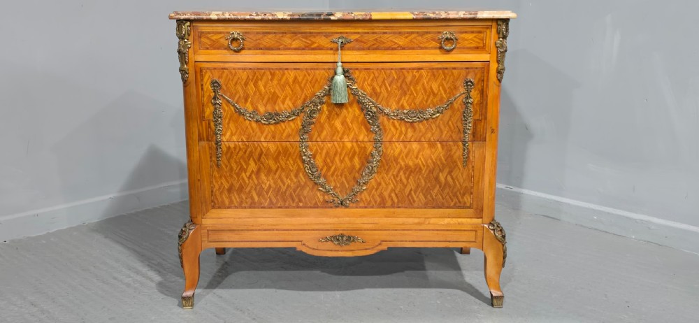 A French satinwood commode chest featuring herringbone parquetry veneer, a variegated marble top, and decorative gilt-bronze mounts. View II