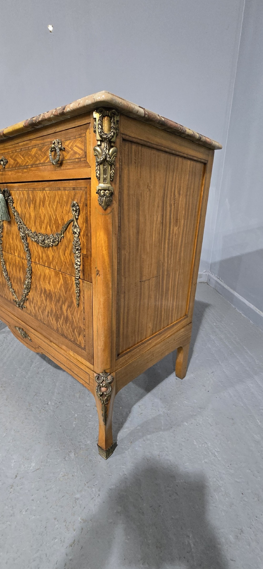 A French satinwood commode chest featuring herringbone parquetry veneer, a variegated marble top, and decorative gilt-bronze mounts. View VI