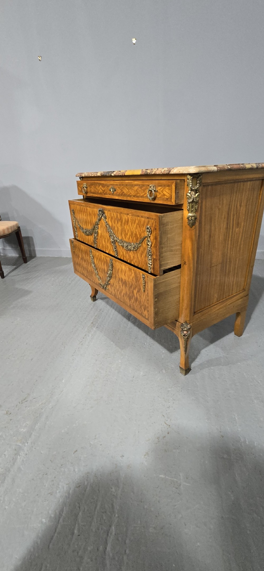 A French satinwood commode chest featuring herringbone parquetry veneer, a variegated marble top, and decorative gilt-bronze mounts. View X