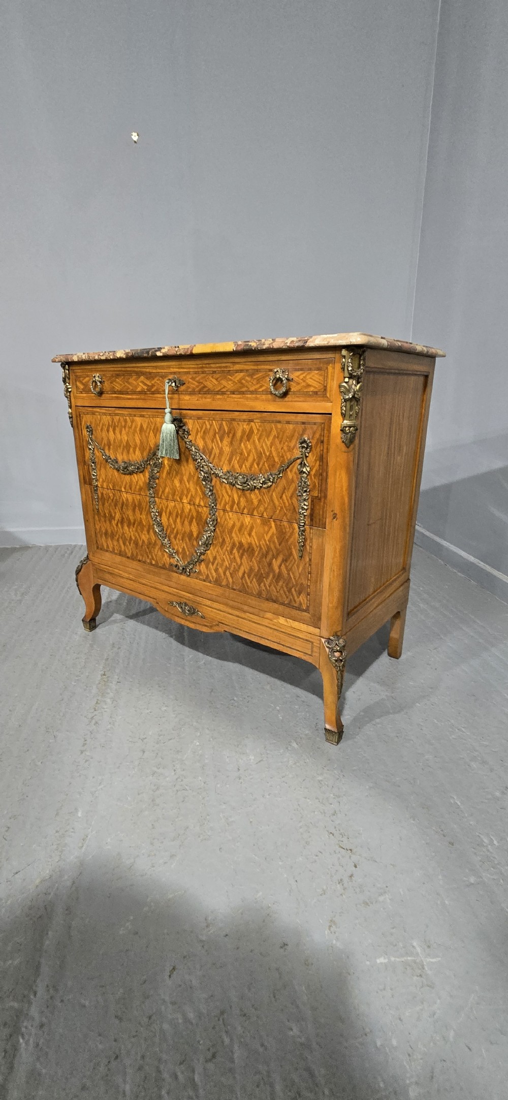 A French satinwood commode chest featuring herringbone parquetry veneer, a variegated marble top, and decorative gilt-bronze mounts. View XVI