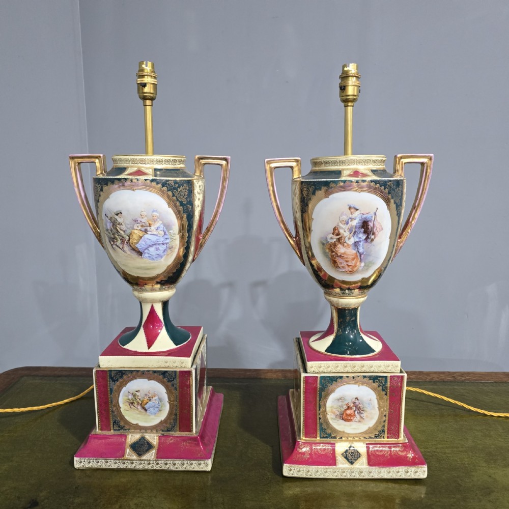 A pair of tall urn-form porcelain table lamps featuring hand-painted classical figural panels, intricate gold gilding, twin handles, and stepped pedestal bases.