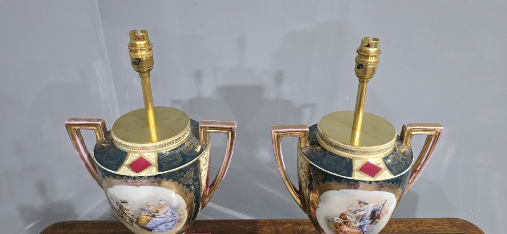 A pair of tall urn-form porcelain table lamps featuring hand-painted classical figural panels, intricate gold gilding, twin handles, and stepped pedestal bases. View VI