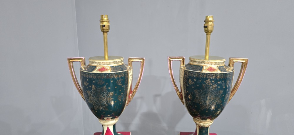 A pair of tall urn-form porcelain table lamps featuring hand-painted classical figural panels, intricate gold gilding, twin handles, and stepped pedestal bases. View IX