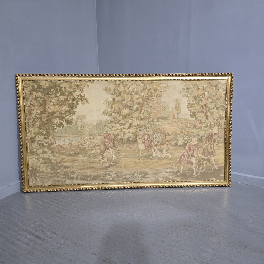 A large horizontal framed tapestry depicting an 18th-century style French hunting scene with horse riders, hounds, and a woodland landscape within a gilded beaded frame.