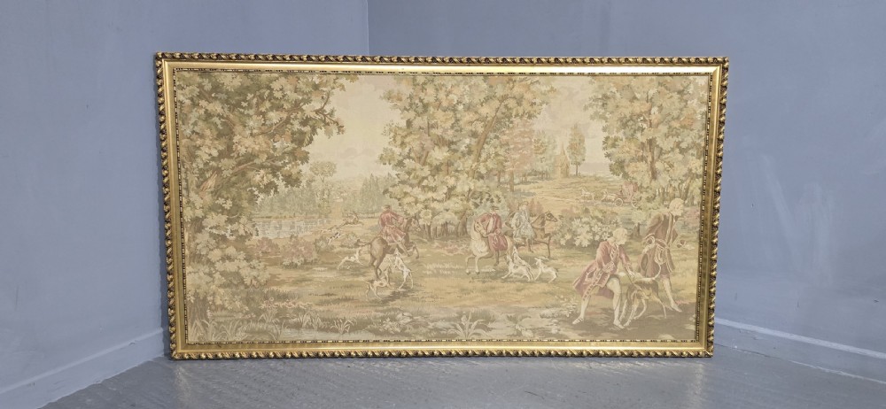 A large horizontal framed tapestry depicting an 18th-century style French hunting scene with horse riders, hounds, and a woodland landscape within a gilded beaded frame. View II