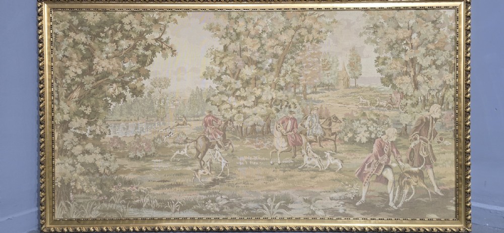 A large horizontal framed tapestry depicting an 18th-century style French hunting scene with horse riders, hounds, and a woodland landscape within a gilded beaded frame. View III