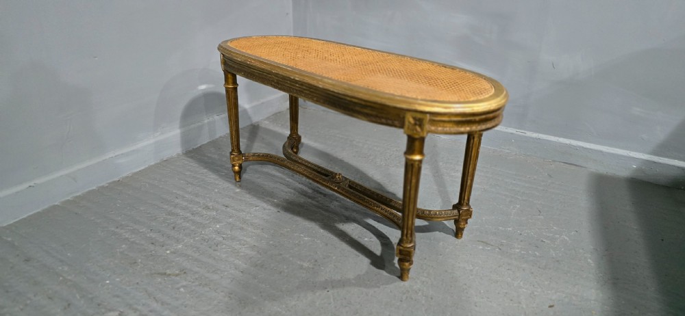 An oval French Louis XVI style duet stool featuring a carved giltwood frame with fluted legs and a gold silk satin upholstered cushion. View VII
