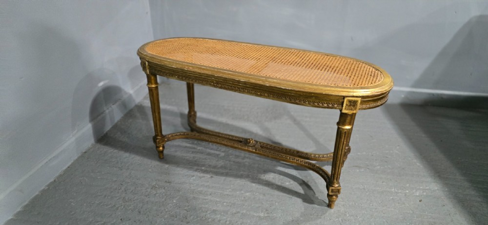 An oval French Louis XVI style duet stool featuring a carved giltwood frame with fluted legs and a gold silk satin upholstered cushion. View VIII