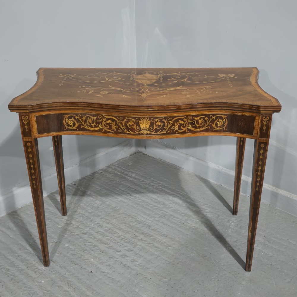 An antique mahogany card table featuring detailed marquetry inlay, a serpentine front, an ornamental urn motif, and square tapered legs.