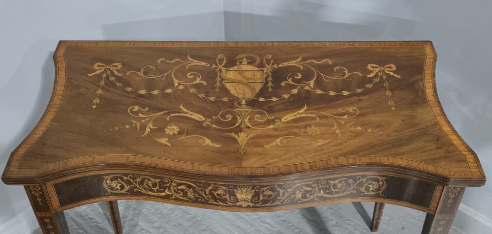 An antique mahogany card table featuring detailed marquetry inlay, a serpentine front, an ornamental urn motif, and square tapered legs. View II