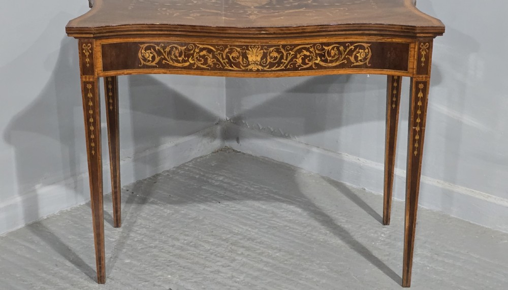 An antique mahogany card table featuring detailed marquetry inlay, a serpentine front, an ornamental urn motif, and square tapered legs. View VI