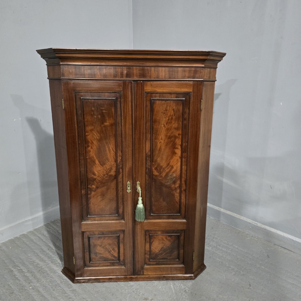 A floor-standing Regency mahogany corner cupboard featuring double panelled doors with flame mahogany veneers and central brass escutcheons.